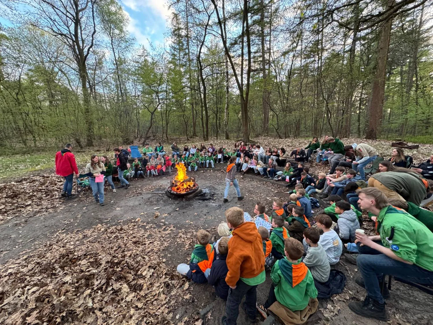 Grote kring van scouts van alle leeftijden rond een kampvuur in het bos