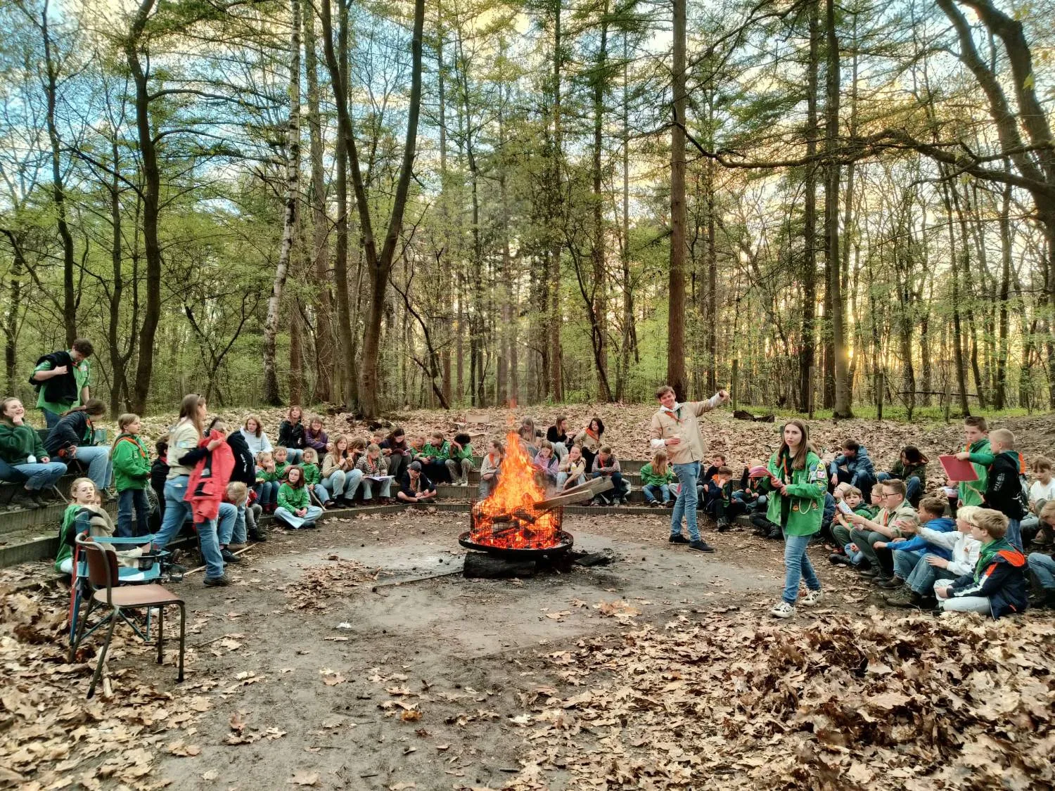 Scouts zitten op de tribune van de kampvuurkuil terwijl de leiding bij het vuur een spel leidt