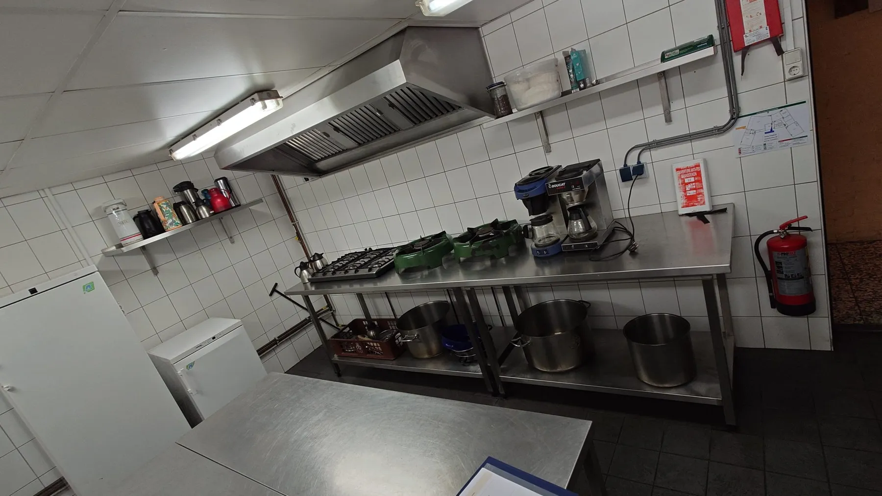 Keuken met RVS werktafels, gasbranders, afzuigkap, grote pannen en een koelkast
